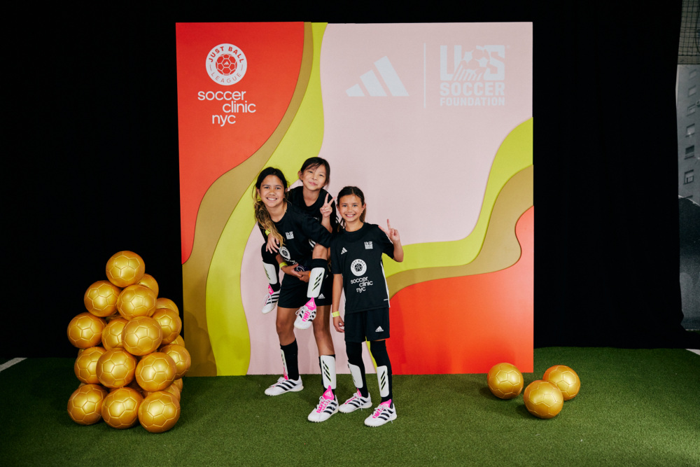 Three girls pose in front of a soccer backdrop at the NYC soccer clinic